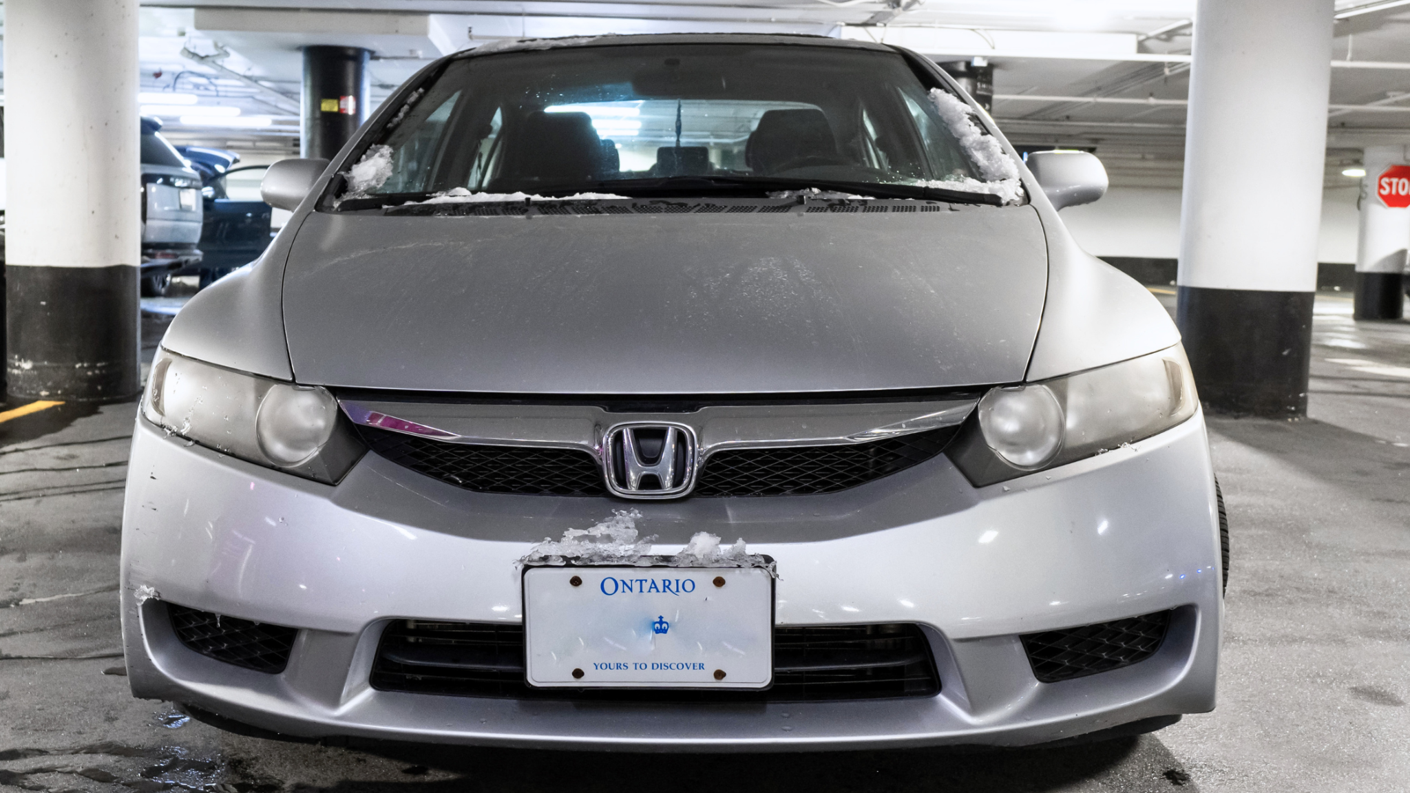 Front view of a silver Honda Civic with salt residue and snow buildup on the hood and windshield, parked inside an underground garage with visible Ontario license plate.