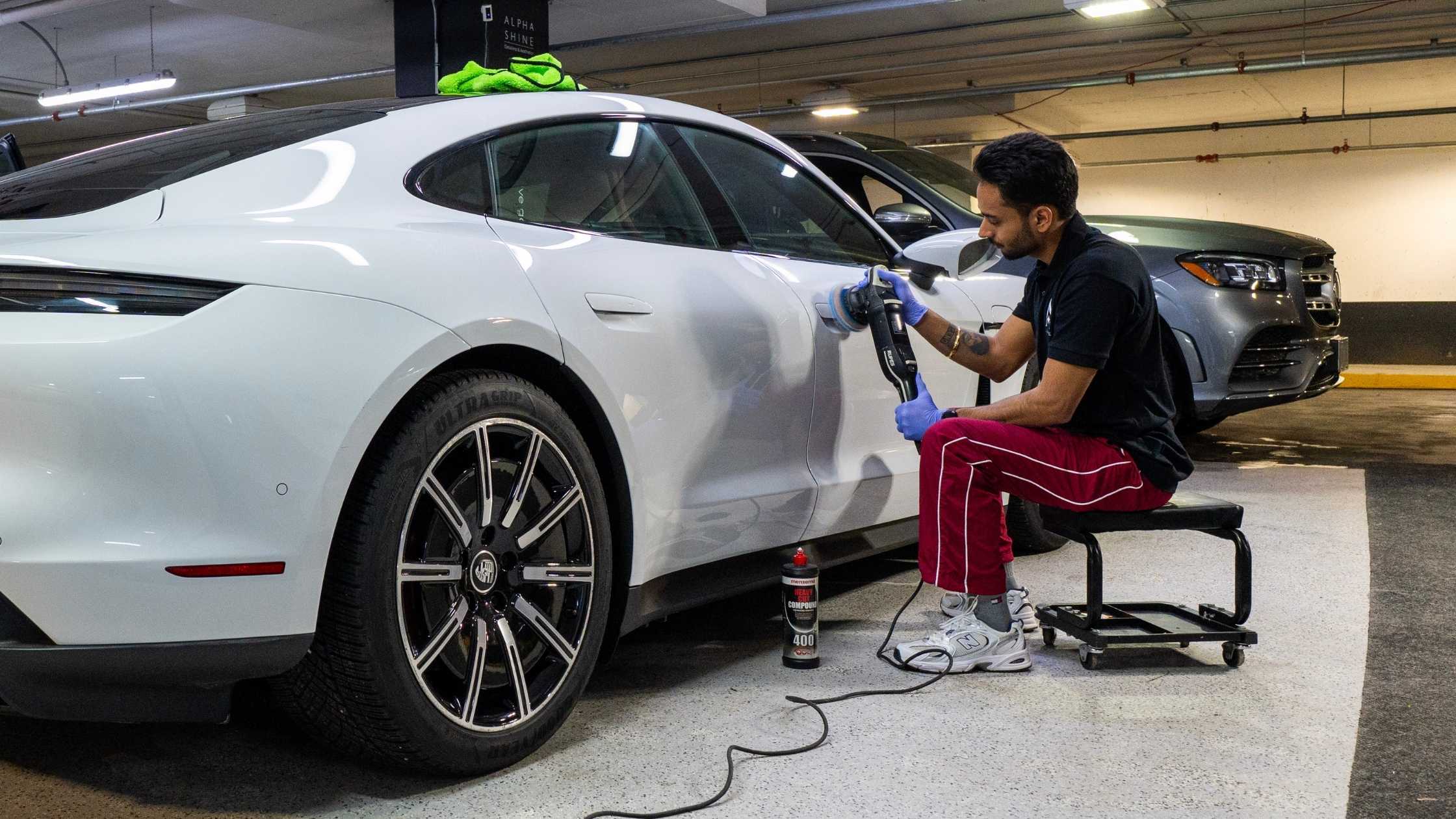 Detailer polishing the side panel of a white car using a machine polisher, seated on a rolling stool in a garage.