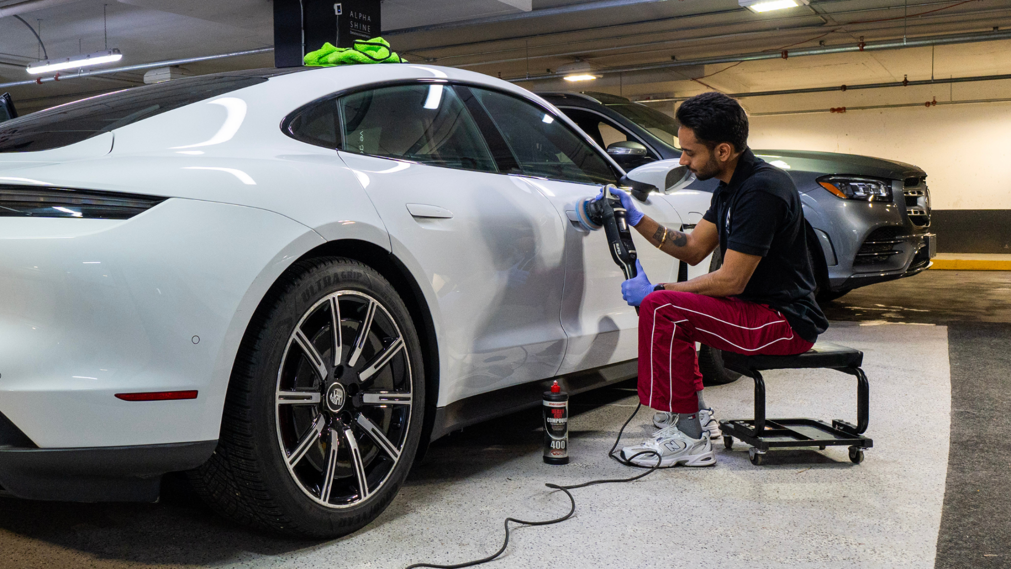Detailer polishing the side panel of a white car using a machine polisher, seated on a rolling stool in a garage.