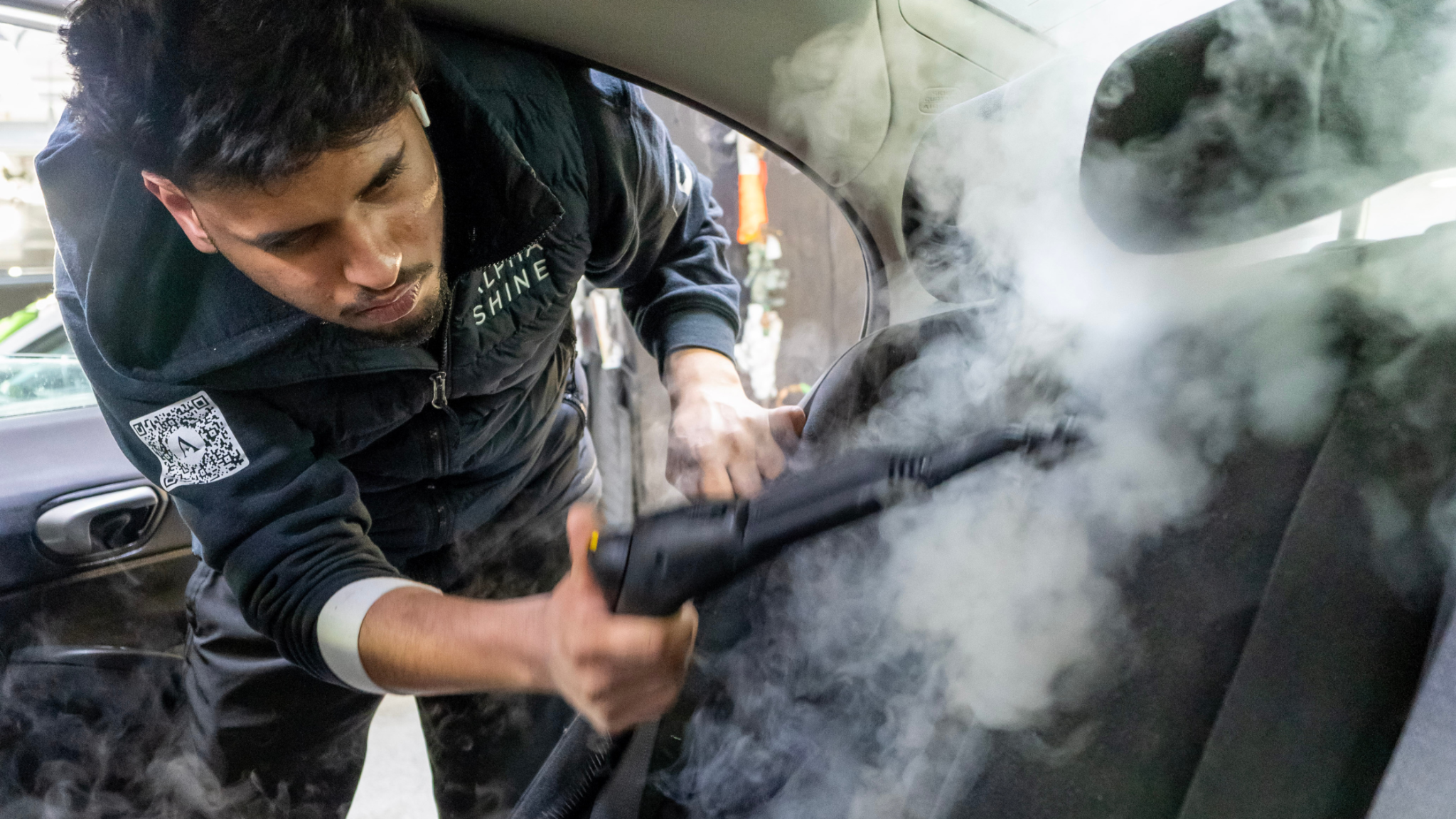 ALPHASHINE detailer using a steam cleaner to deep clean a car seat, with visible steam lifting dirt from fabric upholstery inside a vehicle.