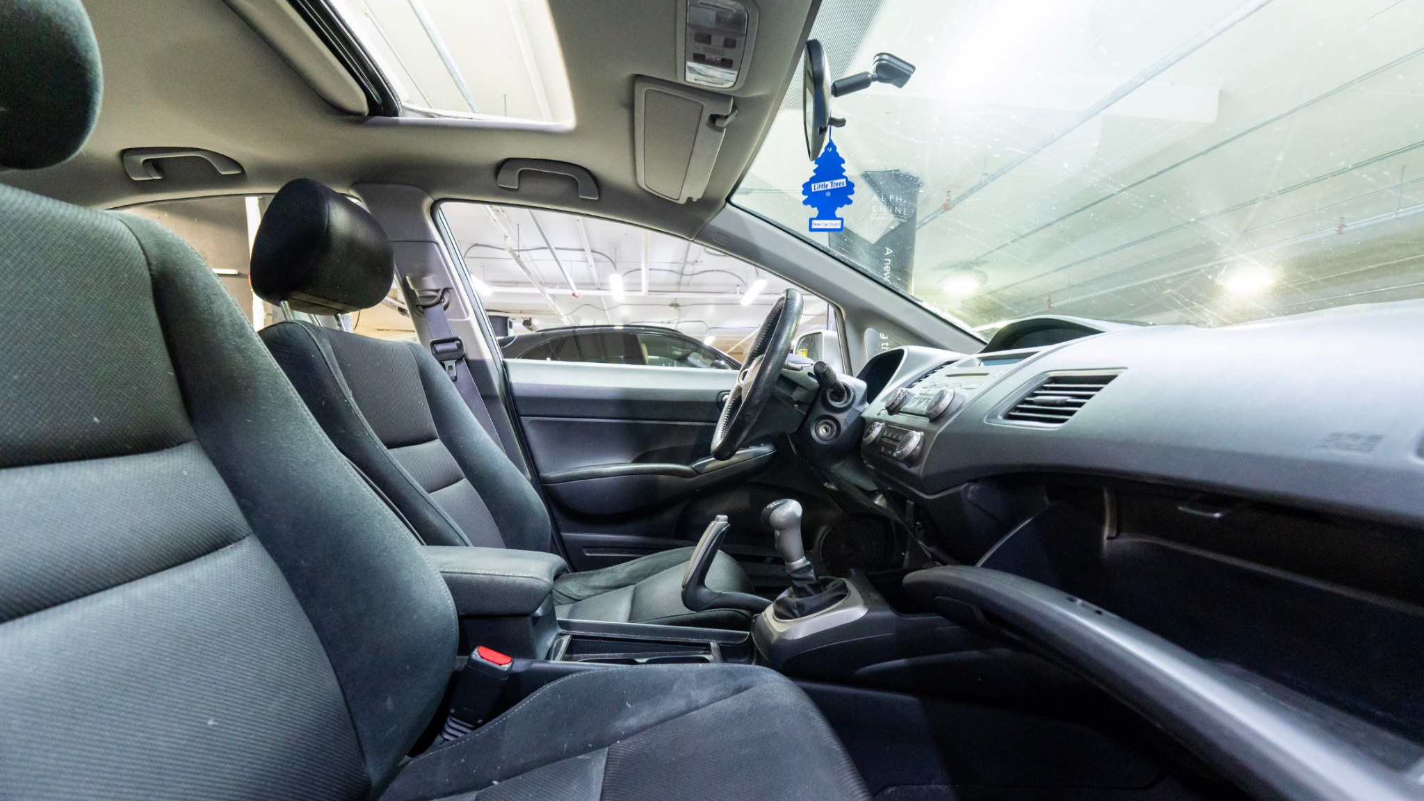 Clean car interior with fabric seats, center console, and manual gear shift, parked inside an underground garage with bright overhead lighting.