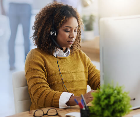 Woman with curly hair wearing a headset and a mustard sweater, typing at a desk in front of a computer.