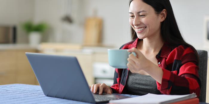 Smiling woman sitting at a table with a laptop, holding a coffee mug while working in a bright home kitchen.