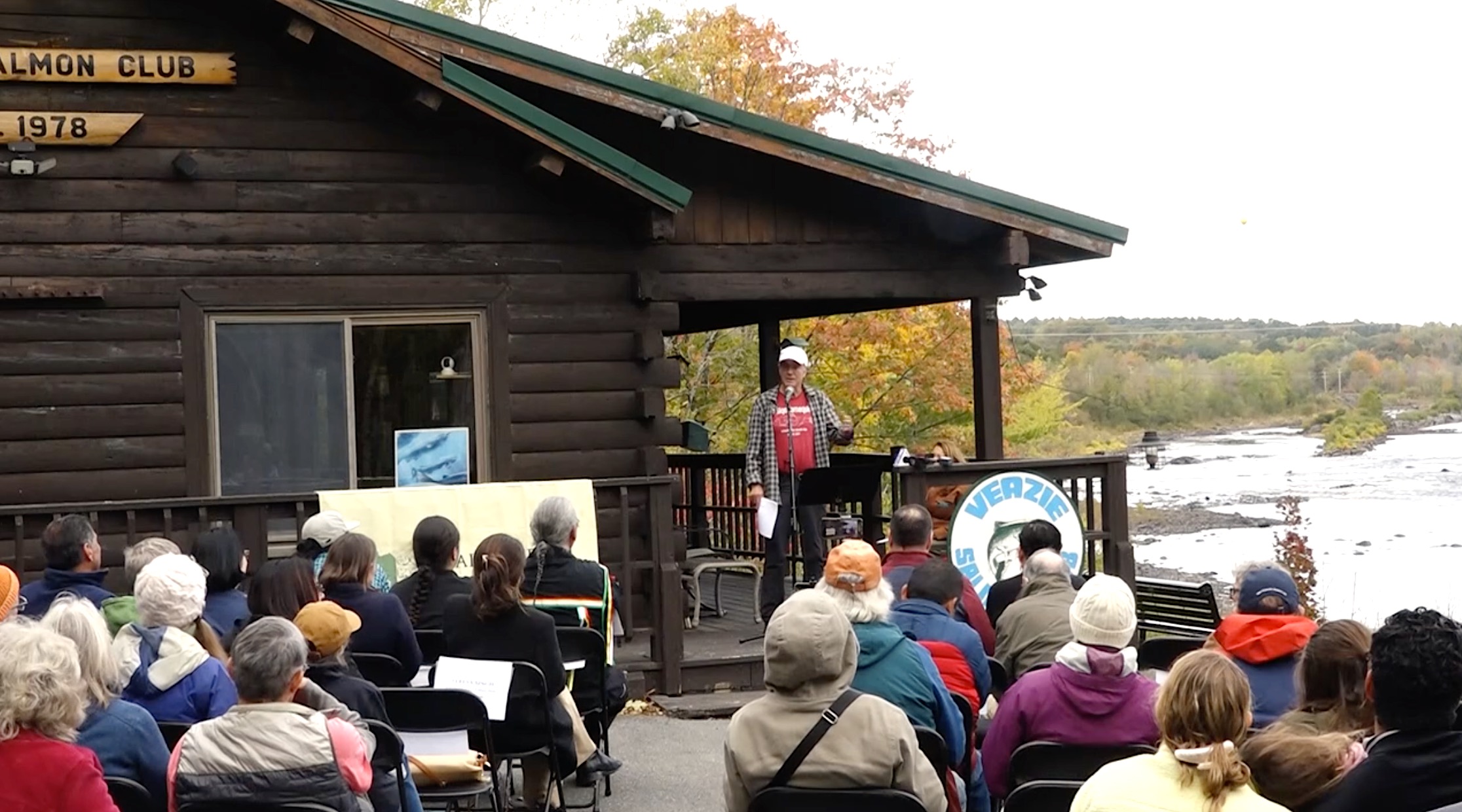 Speaker standing on a wooden porch in front of a small building addressing an outdoor audience seated near a river with autumn trees in the background.