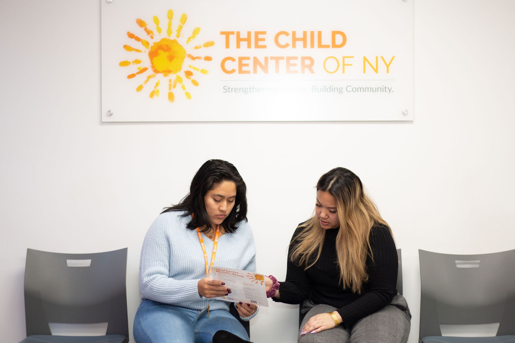 Two women sitting down on gray chairs reading a piece of paper in front of a The Child Center of NY banner