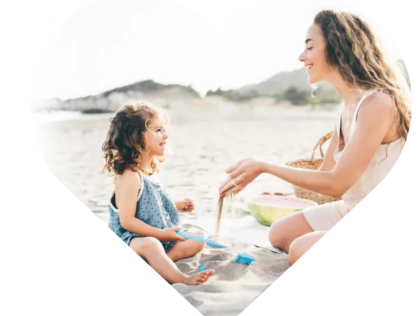 Au pair and young girl playing with sand and beach toys, smiling at each other on a sandy beach.