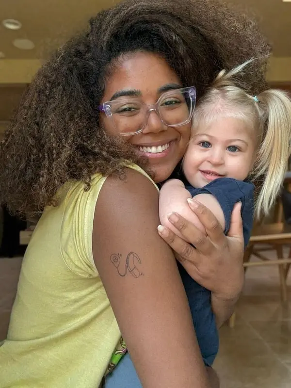 Smiling au pair with glasses and curly hair hugging a young blonde girl with pigtails indoors.