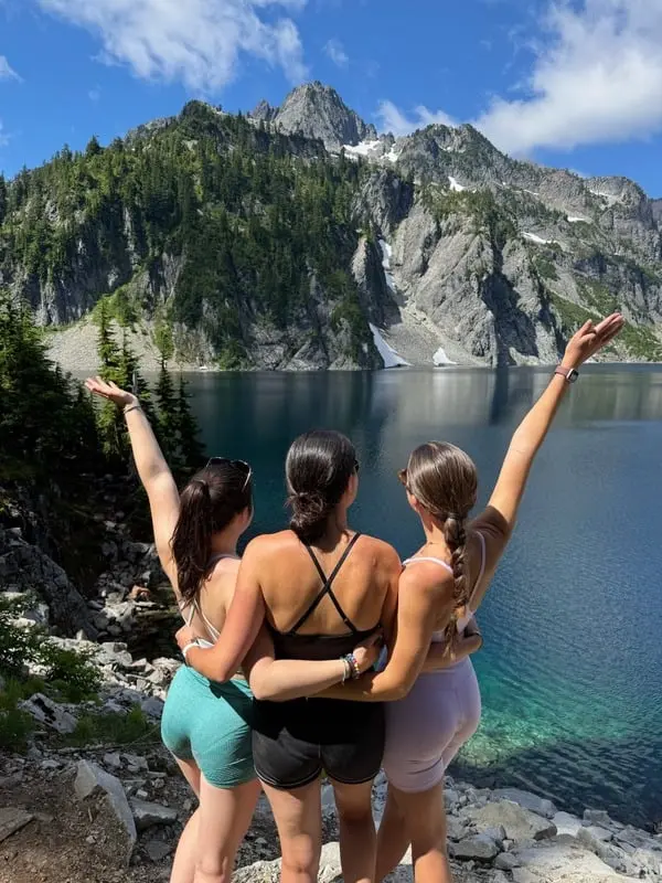 3 au pairs standing with arms around each other, facing a mountain lake and rocky, tree-covered peaks under a blue sky.