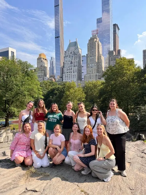 Group of au pairs posing on a large rock with city skyscrapers and green trees in the background under a blue sky.