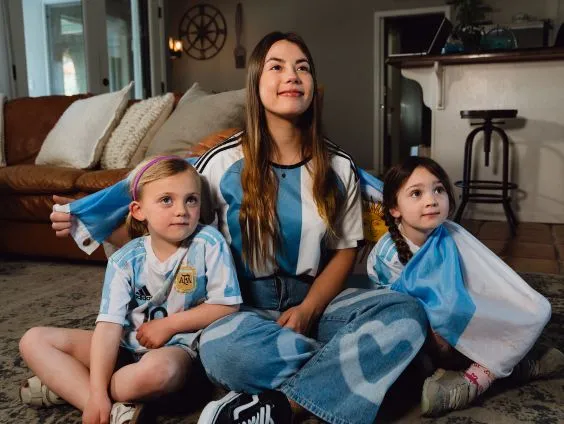 Three smiling girls sitting on a carpet in a living room, two holding Argentine flags and wearing Argentina soccer jerseys.