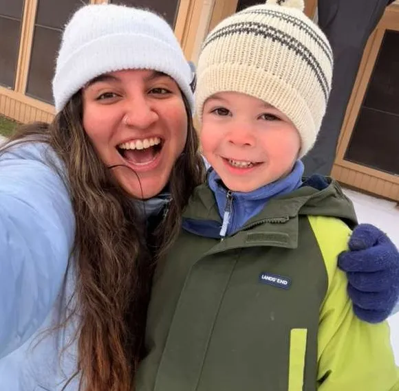 Woman and child in winter coats and hats smiling outdoors with snow on the ground.