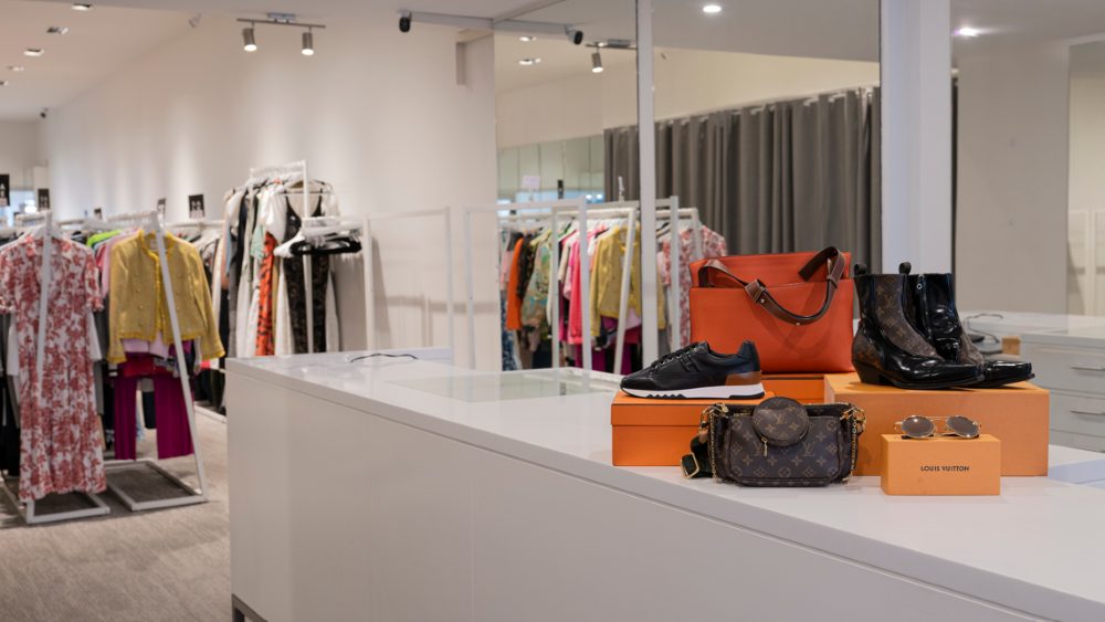 Preloved Louis Vuitton bag, boots, sneakers and sunglasses on the counter of a VENLA store, with racks of designer fashion behind
