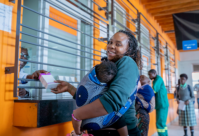 Florine, from Zambia, with her baby in her arms smiles to camera as she receives medication at her local pharmacy