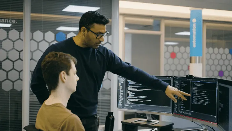 Two men collaborating in an office, one seated and one standing pointing at code on dual computer monitors.