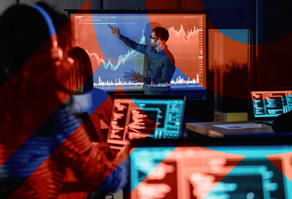 Man pointing to a large screen displaying stock market charts while people in foreground work on laptops with code and data.