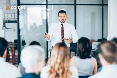 Man in white shirt and tie speaking to an audience in a modern meeting room with glass walls.