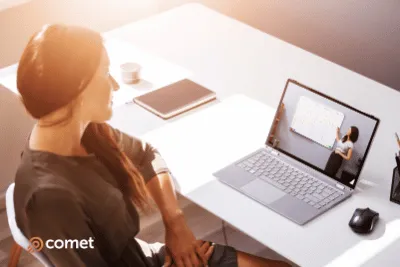 Woman with long dark hair sitting at a white desk, watching a laptop screen showing a person presenting in front of a board.