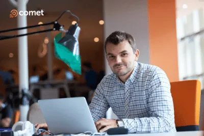 Smiling man in a checkered shirt working on a laptop at a desk with a green-shaded desk lamp and orange chair.