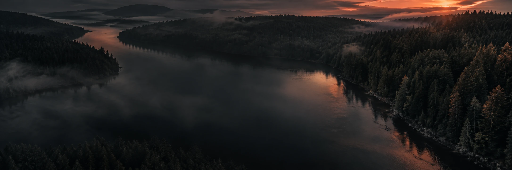 An aerial view of a forested loch at dawn with mist lying across the water