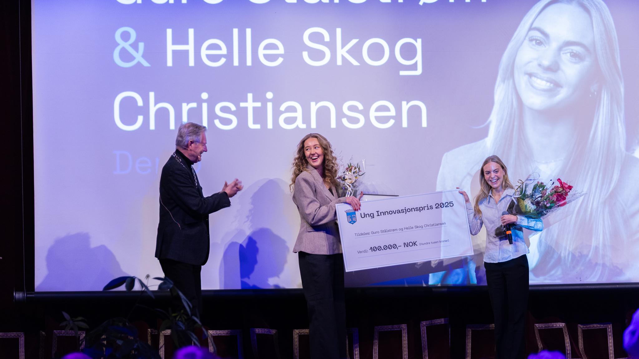 Bilde av de glade vinnerne Helle Skog Christiansen og Guro Stålstrøm som får gavesjekk, diplom og blomster av varaordfører Erling Moe på scenen på Studentersamfundet. 