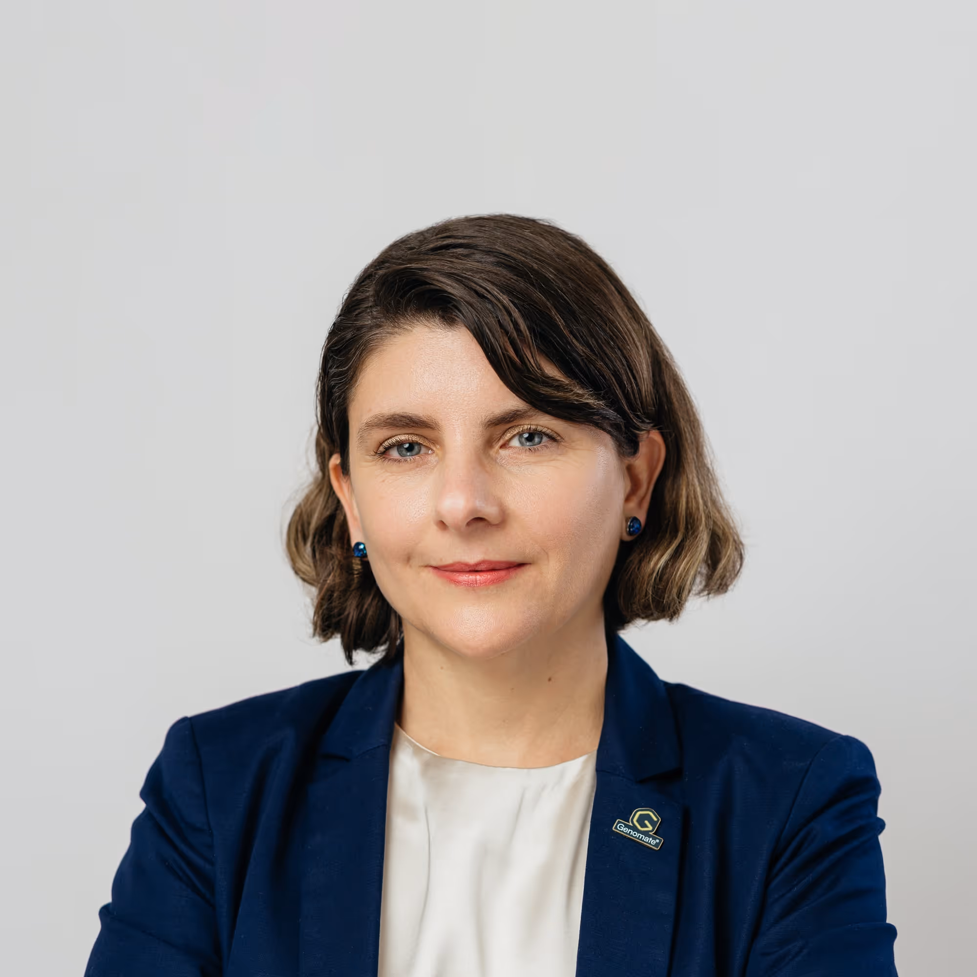 Orsolya Peter with short brown hair wearing a navy blazer and white top against a plain gray background.