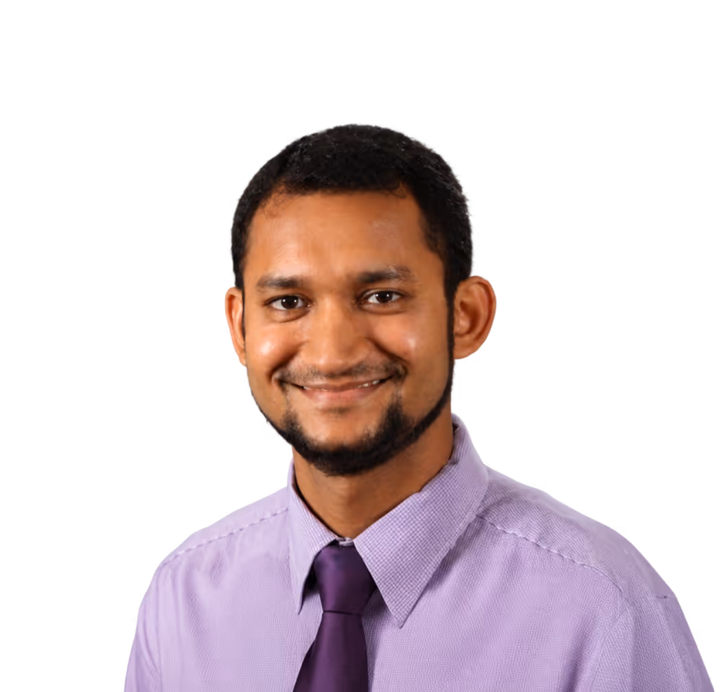 Man smiling, wearing a light purple shirt and a dark purple tie against a plain gray background.