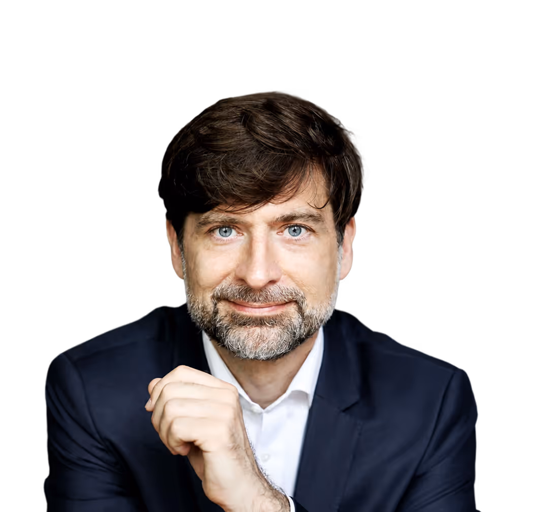 Man with blue eyes and beard wearing dark suit and white shirt, smiling with hand near chin.