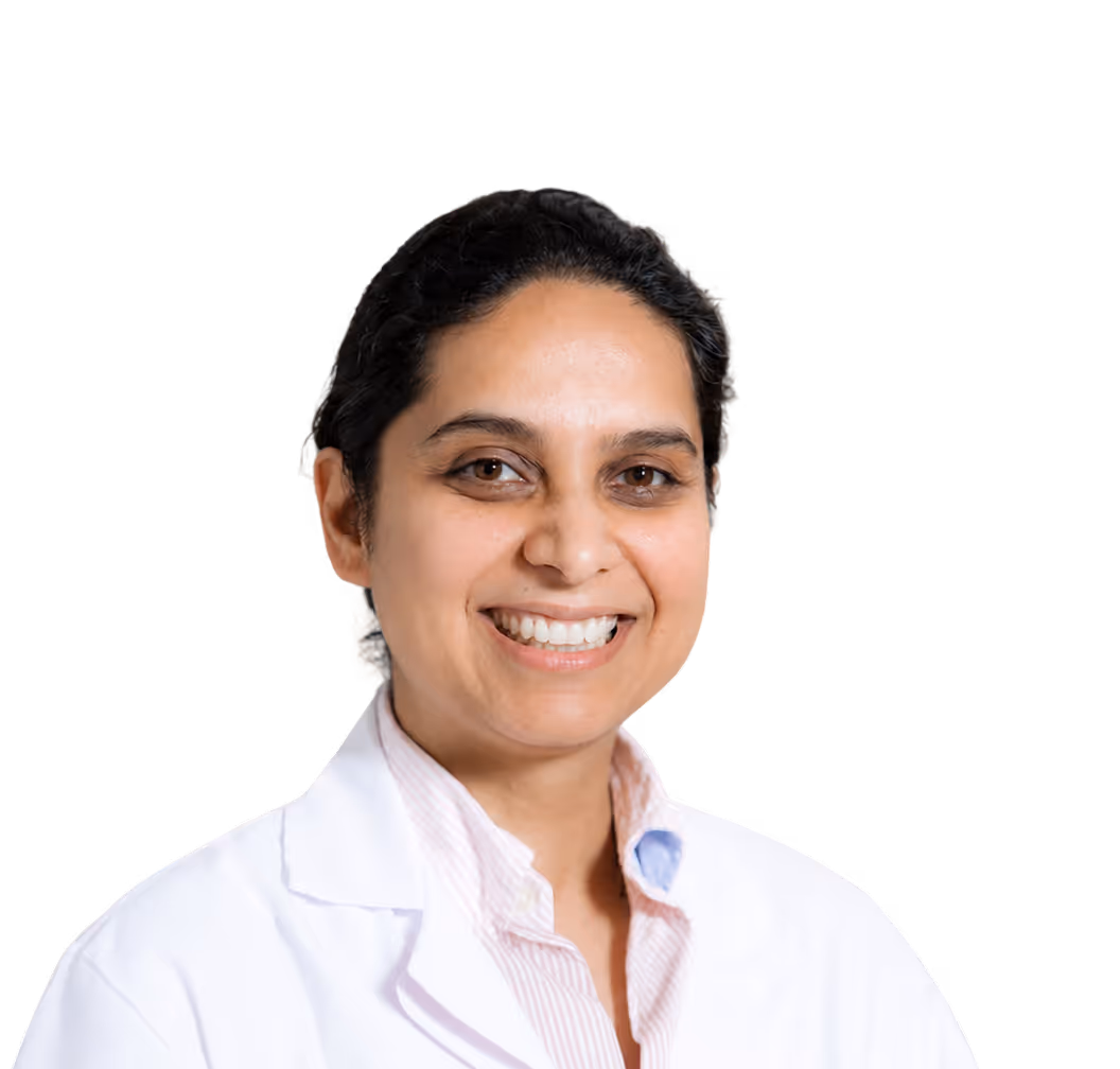 Smiling woman with dark hair wearing a white medical coat against a plain light background.