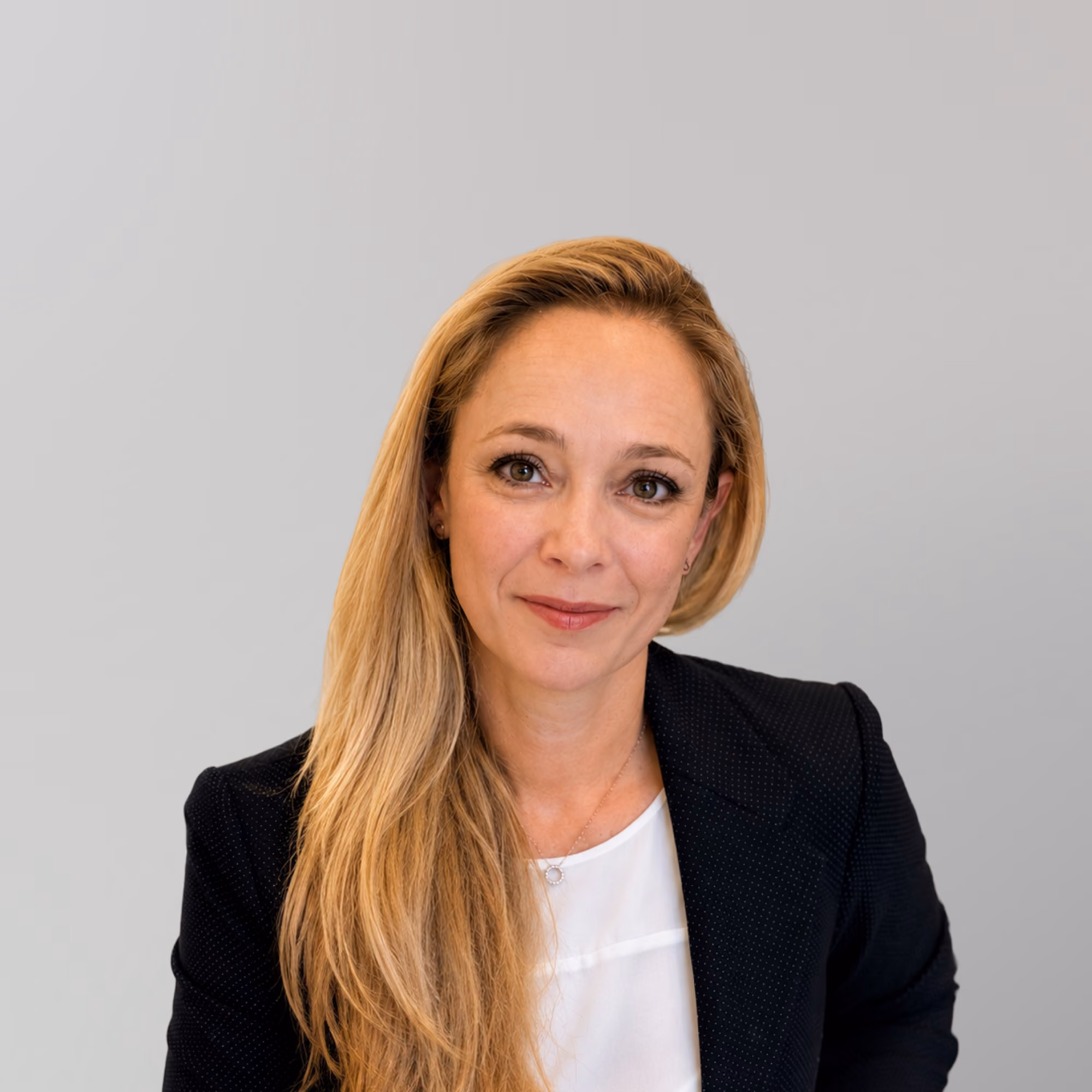 Smiling Petra Felszghy with long blonde hair wearing a black blazer and white top against a gray background.