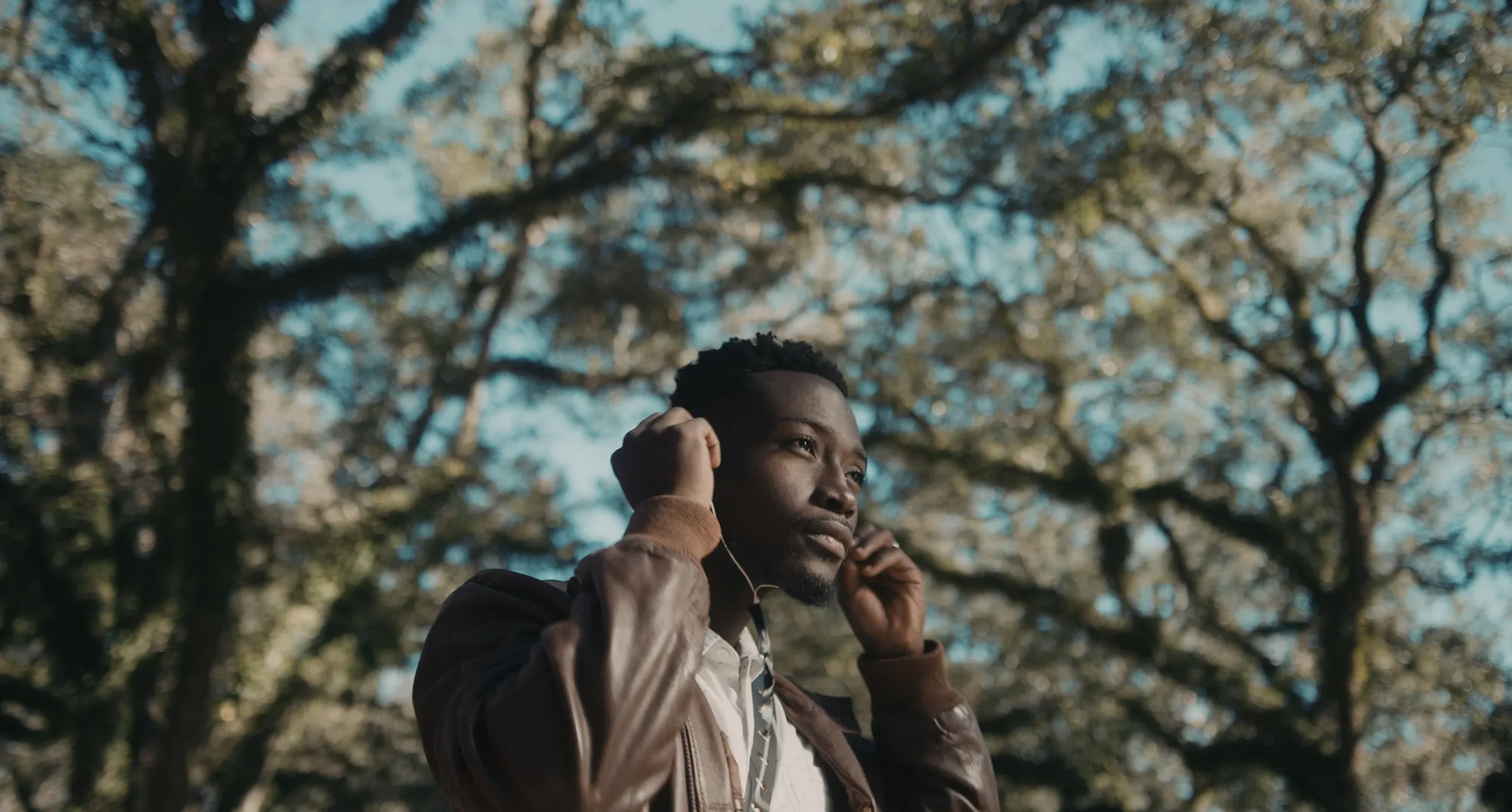 17 Year Locust film still showing the young Haitian man Renee putting in earbuds, green trees surrounding him. 