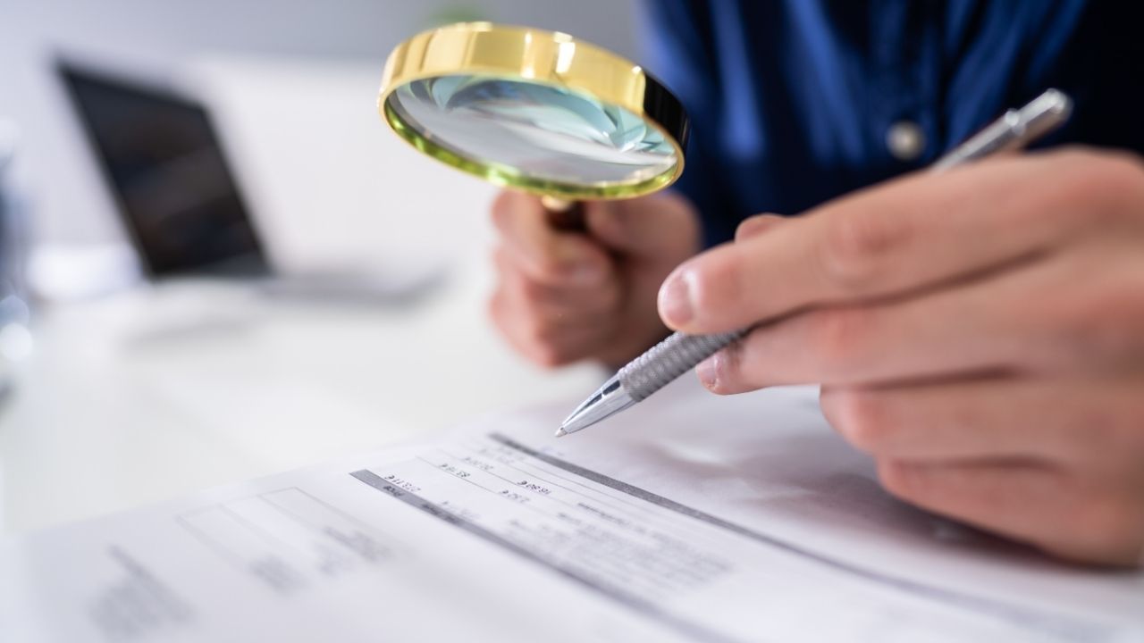 Close-up Of A Businessperson's Hand Looking At Receipts Through Magnifying Glass At Workplace
