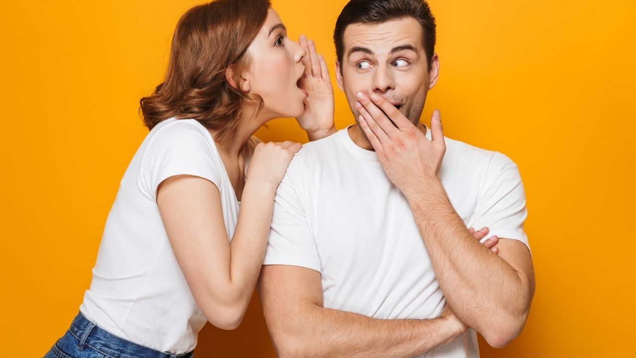 Woman whispering a secret to a surprised man covering his mouth, both against an orange background