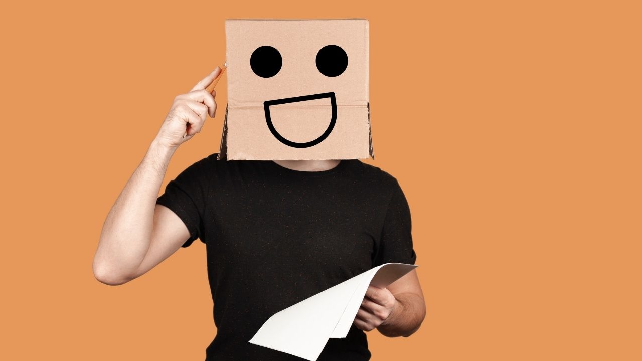 Person with a smiley-face cardboard box over their head taps their temple with a pencil while holding papers, on an orange background.