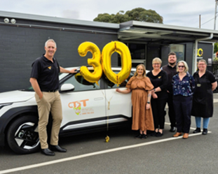 A group of people standing near a car holding a gold 30 sign