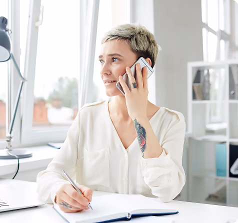 Person with short hair on phone call writing in notebook at desk near window.
