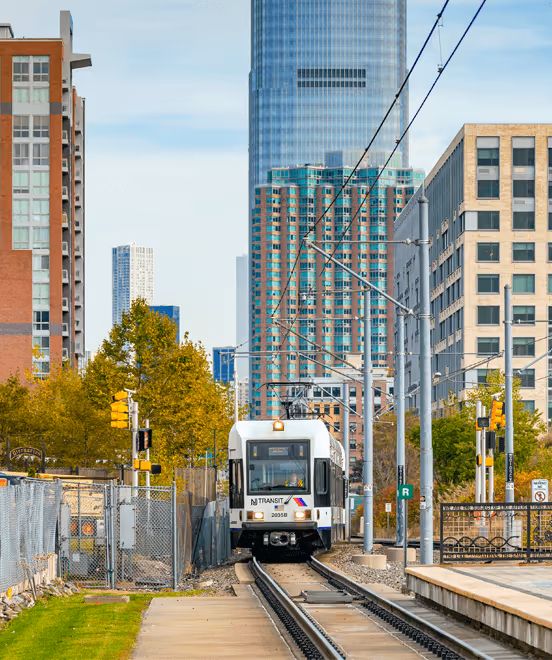 NJ Transit light rail train traveling on tracks through urban area with tall buildings and trees in the background.