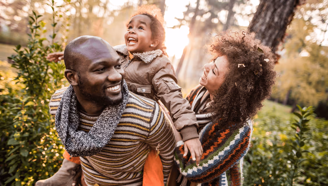 Smiling family of three outdoors in autumn, with father giving daughter a piggyback and mother holding daughter's hand.