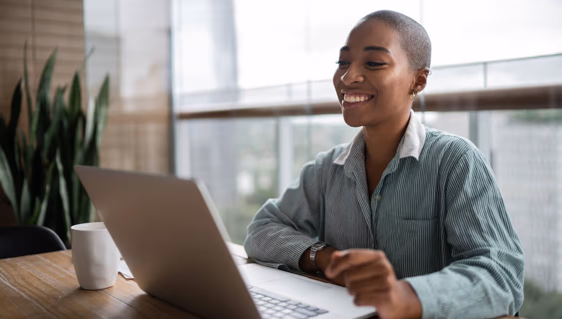 Smiling woman with short hair sitting at a wooden table and using a laptop near a window.