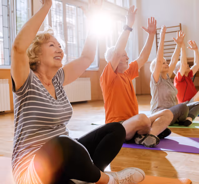 Group of older adults sitting cross-legged on yoga mats indoors, raising their arms during a yoga class, with sunlight streaming through the windows.