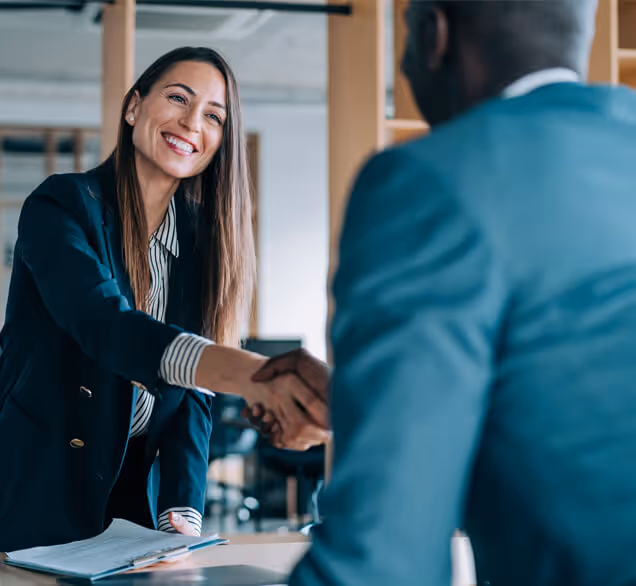 Smiling woman in a blazer shaking hands with a man in a suit in an office setting.