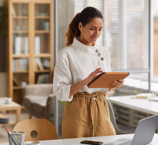 Woman standing in a modern office using a tablet, smiling with wireless earbuds in.