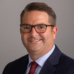 Smiling man with glasses wearing a navy suit, light blue shirt, and red striped tie against a gray background.