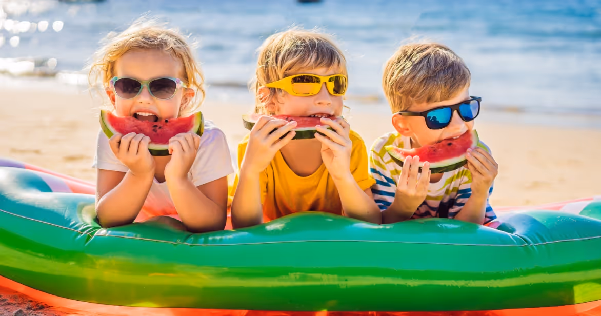 Three children in sunglasses eating watermelon on a beach during summer – fostering in Ireland summer event July 29th.
