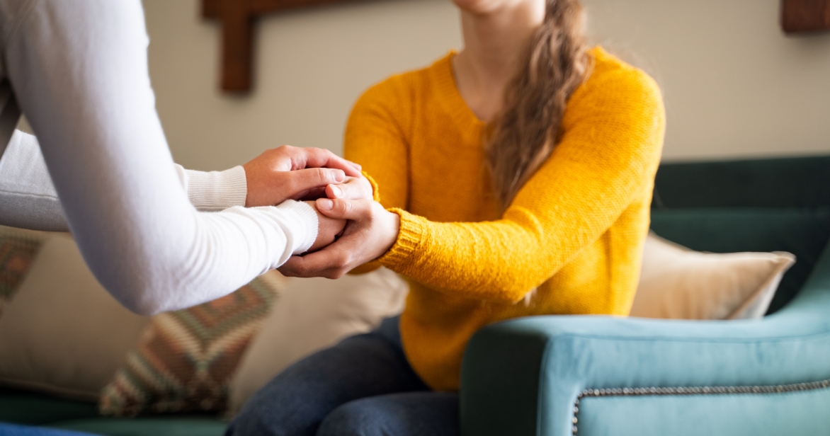 Close-up of two women holding hands in support, seated on a sofa, representing how Nicola found strength in support with Origins