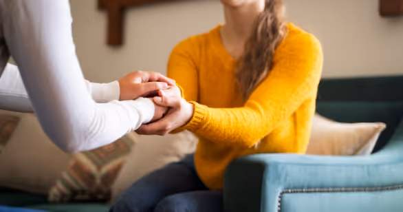 Close-up of two women holding hands in support, seated on a sofa, representing how Nicola found strength in support with Origins