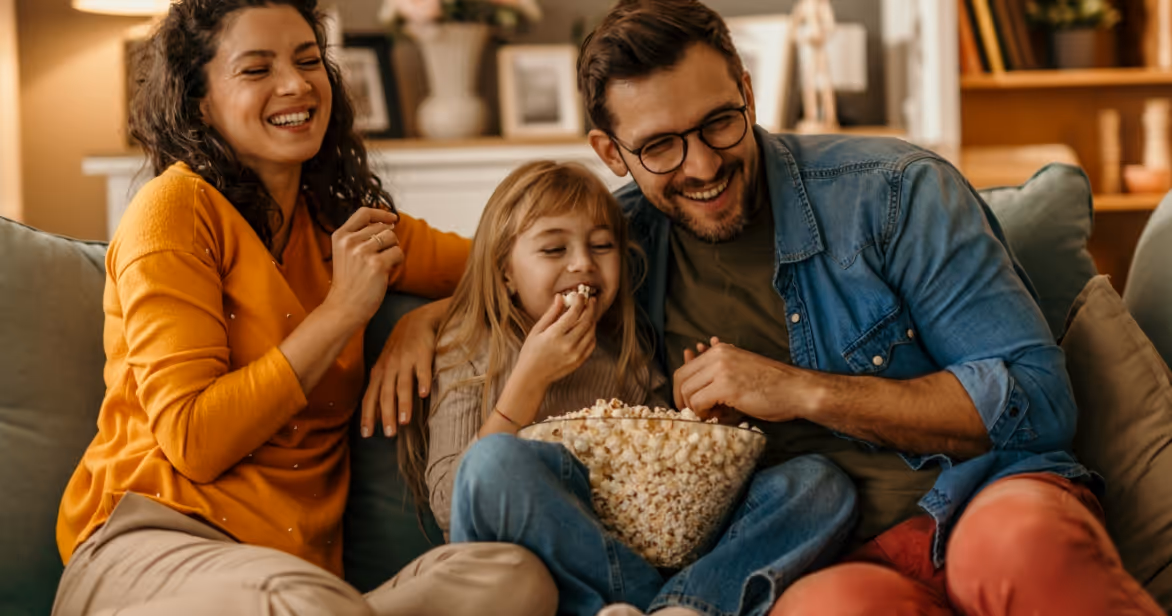Foster family laughing together on the sofa, sharing popcorn during quality time at home.