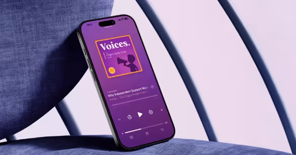 An iPhone displaying the Voices – The Origins Foster Care Podcast on its screen, resting against a blue fabric chair. The podcast artwork features a purple background with a child holding a megaphone and the Origins Foster Care logo in the corner.