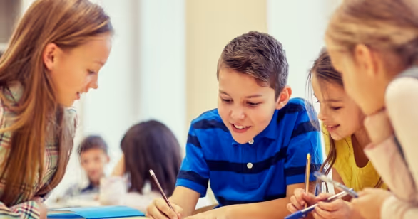 A smiling young boy and three girls sitting together at a classroom table writing in notebooks, representing the stability and educational support needed when growing up in foster care.