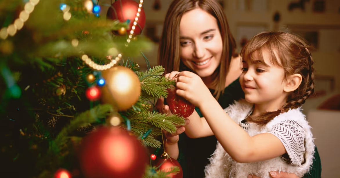 A smiling young girl with brown hair and pigtails decorates a Christmas tree with the help of her foster carer, who gently supports her. The tree is adorned with red and gold baubles and twinkling lights, capturing a warm, festive moment of connection and belonging.
