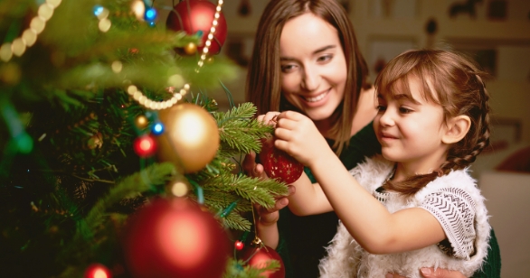 A smiling young girl with brown hair and pigtails decorates a Christmas tree with the help of her foster carer, who gently supports her. The tree is adorned with red and gold baubles and twinkling lights, capturing a warm, festive moment of connection and belonging.